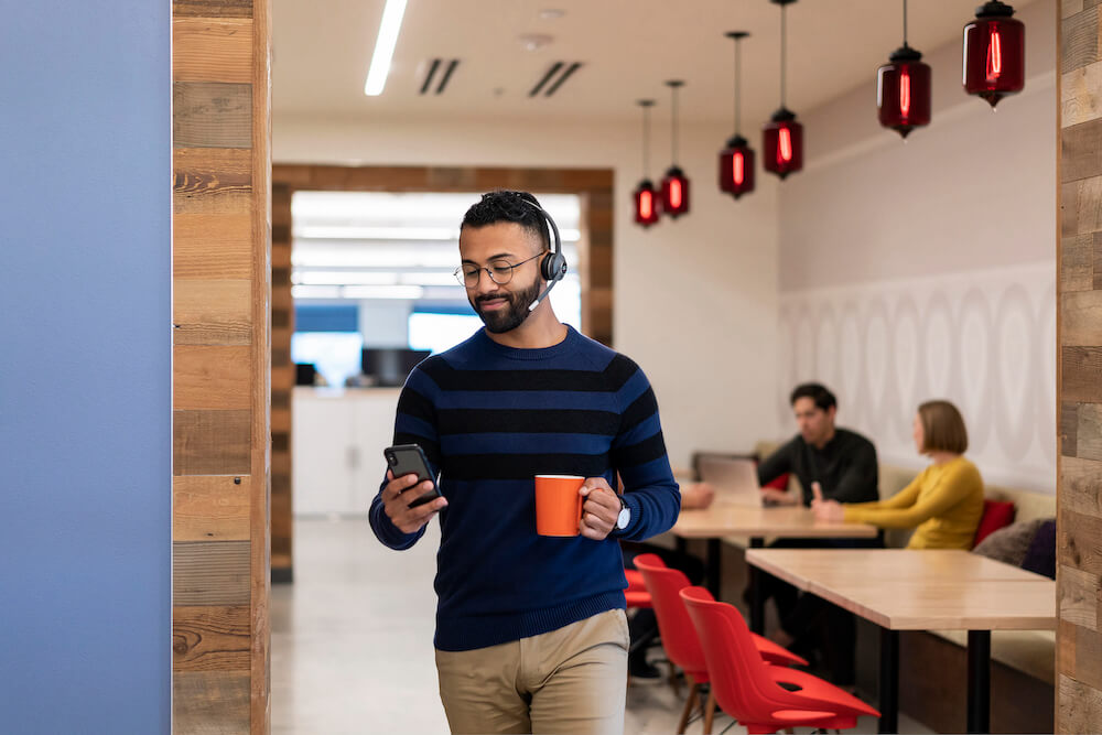 Ein Mann läuft mit einem Kaffee und seinem Mobiltelefon in der Hand durch das Büro, während er das bequeme Cisco Headset trägt