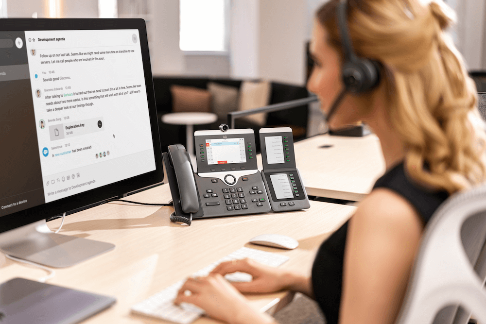 Eine Frau telefoniert &uuml;ber das Cisco IP-Telefon mit Videofunktion und ihr Headset, w&auml;hrend sie Daten in den Computer eintr&auml;gt
