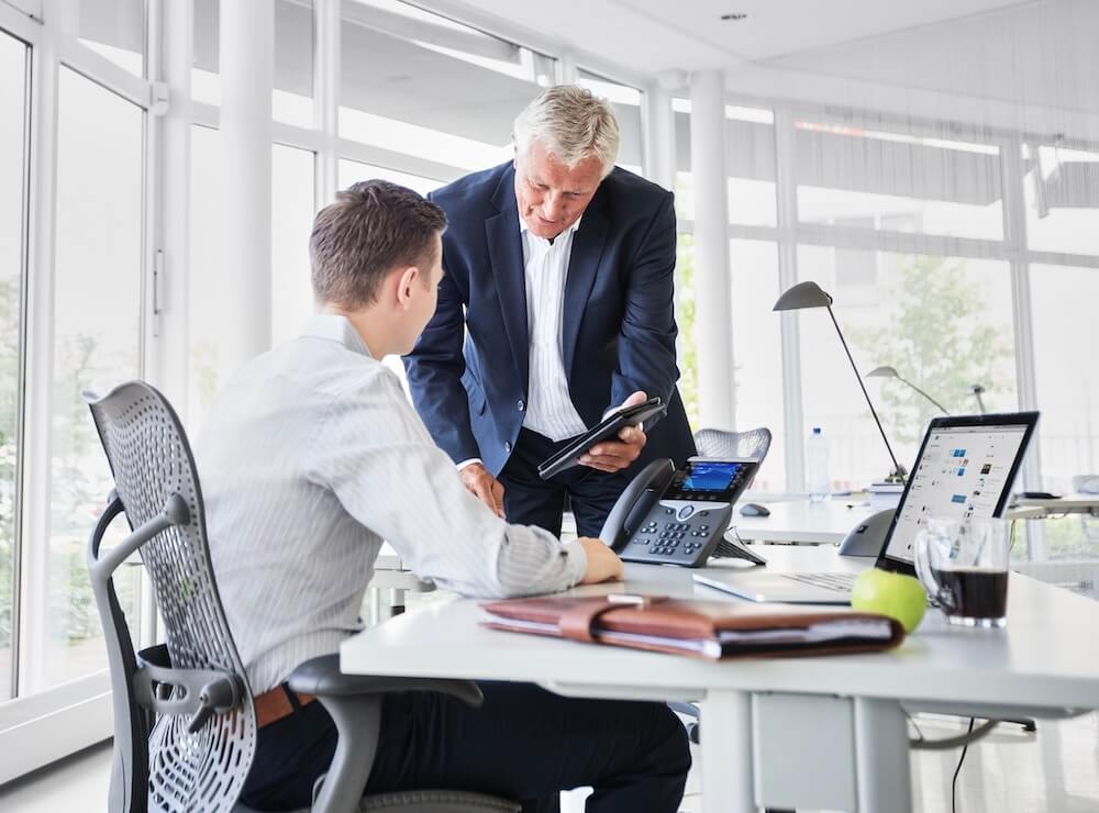 Ein Mann sitzt vor seinem IP-Telefon am Schreibtisch, w&auml;hrend sein Kollege mit ihm spricht
