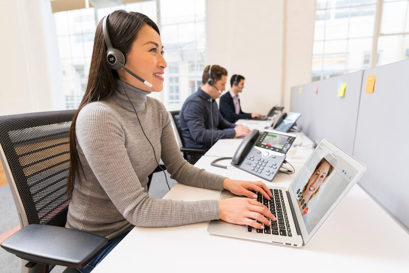 EIne Frau telefoniert im B&uuml;ro &uuml;ber das Headset und ihr IP-Telefon, w&auml;hrend sie den Computer nutzt