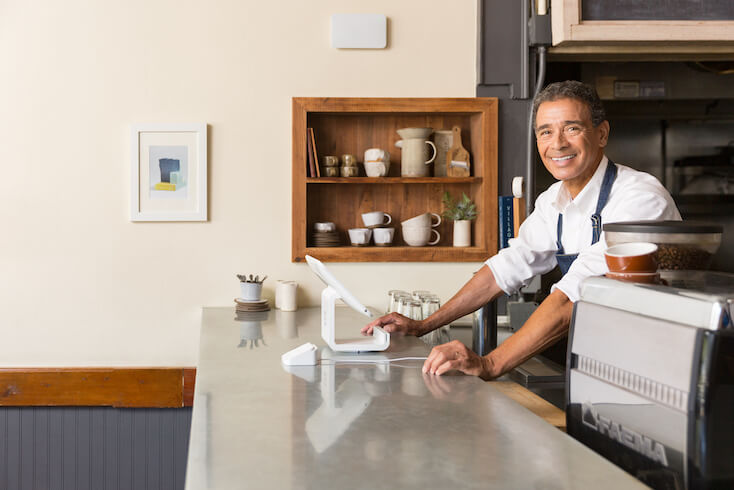 Ein Mann steht neben dem modernen und einfachen Kassensystem seines Cafés, in welchem er seinen Gästen Internetverbindungen bietet